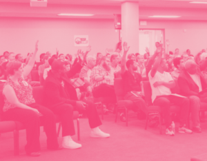People sitting in a large hall raising their hands. It's a casually dressed, mixed-race crowd.