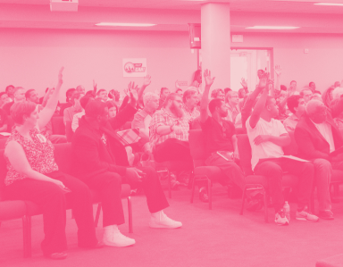 People sitting in a large hall raising their hands. It's a casually dressed, mixed-race crowd.