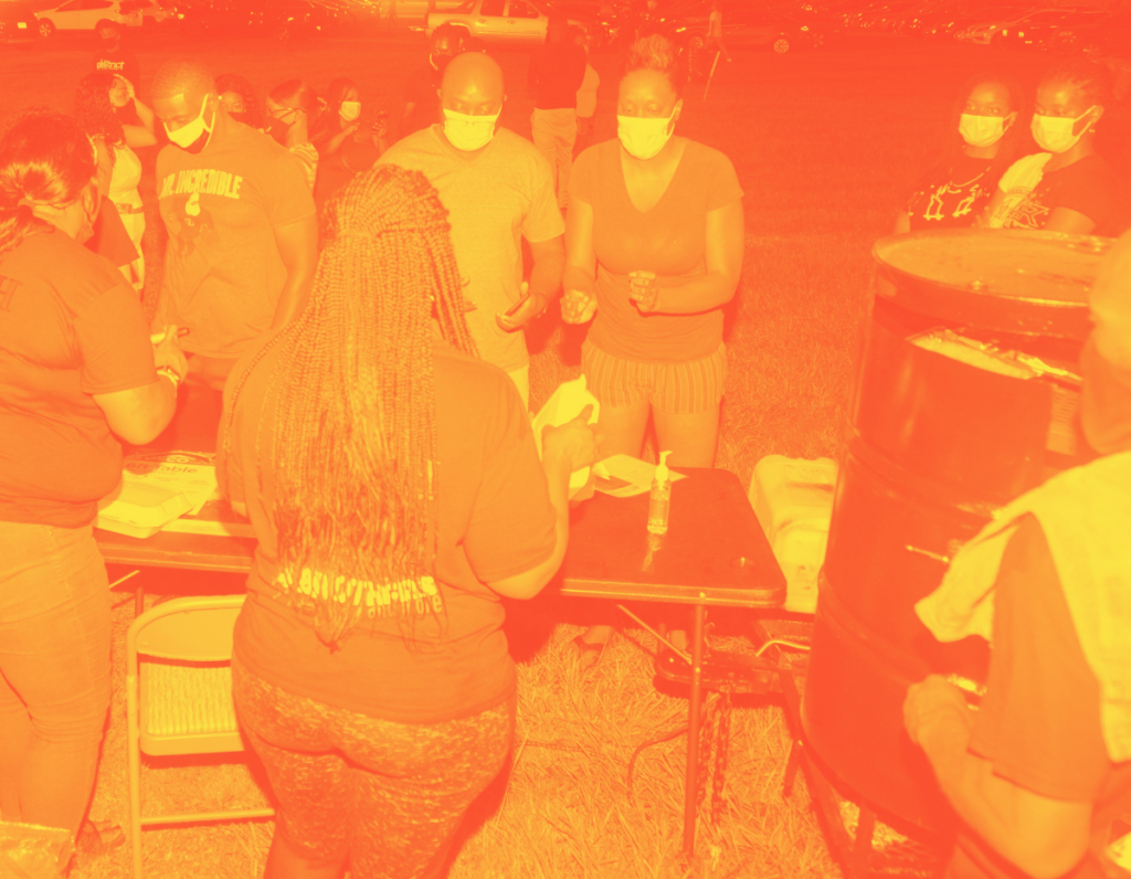 A knot of Black people gather around a folding table. They're masked, dressed for summer. It's dark out.