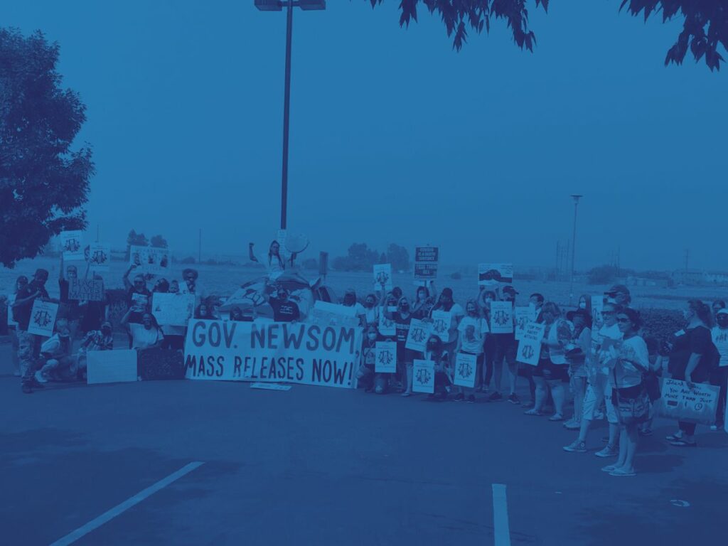 Crowd standing in a semi-circle on a parking lot, with a dusty brown field stretching behind them. Most are women; they’re dressed for summer in shorts and T-shirts. Many hold signs that read, “State negligence is state execution.” Down in front is a big banner reading, “Gov. Newsom, Mass Releases Now.”