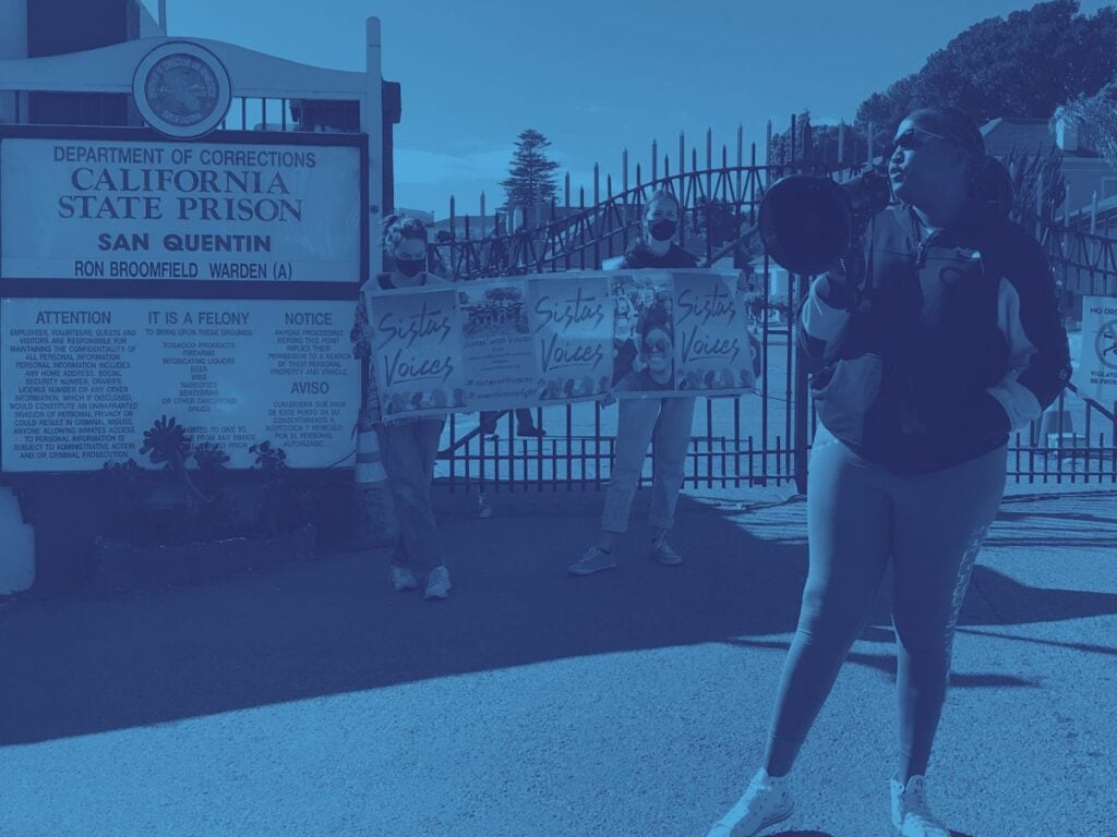 Foreground, Black woman with bullhorn. Behind her, the gates to San Quentin State Prison, with two women both masked, one apparently white and the other apparently API) holding pink “Sistas With Voices” signs standing next to them.