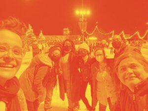 8 people take a group selfie on an ice rink at night with lights hanging around the rink